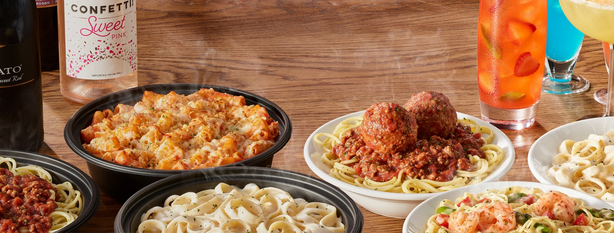 Spaghetti and meatballs, fettuccini Alfredo, baked ziti and bottles of wine on wood table.